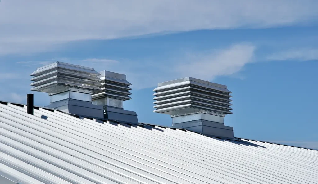A white metal roof with two square-shaped ventilation units protruding from the top, set against a backdrop of a clear blue sky with a few wispy clouds.