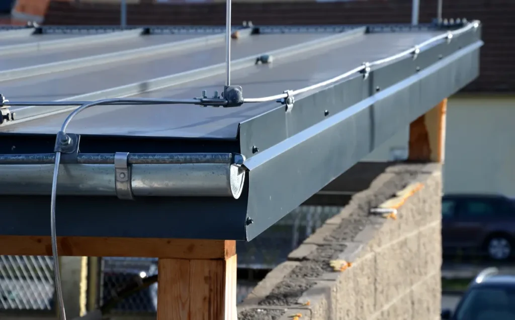 A close-up of a modern solar panel installation on a roof. The panels are mounted on a metal frame atop wooden beams. Cables are visible, suggesting an ongoing or recent installation, with a blurred background of a building.