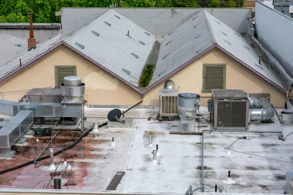 A rooftop with two peaked sections covered in gray shingles, featuring various HVAC units, satellite dishes, and vents. The background shows a tree and surrounding buildings.
