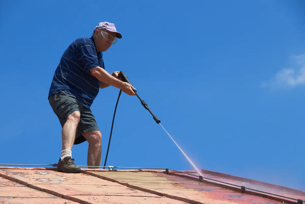 An older man in a striped shirt, shorts, and a cap uses a pressure washer to clean a sloped roof under a clear blue sky.