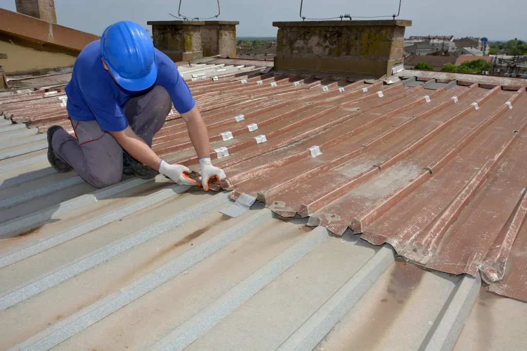 A worker in a blue shirt and helmet repairs a corrugated metal roof. They are using a tool on the rusted edges while kneeling on the roof. The background shows a clear sky and some buildings in the distance.