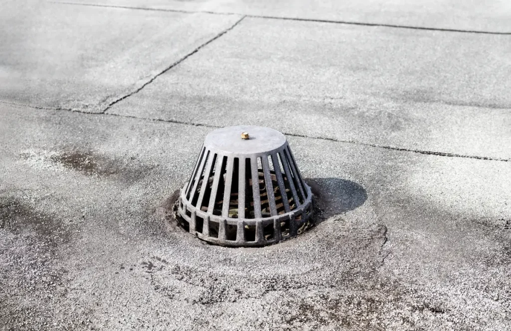 A metal rooftop drain cover is situated on a flat, concrete roof. It features a slatted, conical design to prevent debris from entering. The roof surface shows signs of wear, with visible cracks and patches. Shadows cast around the drain indicate sunlight.