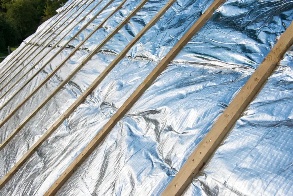Reflective aluminum insulation laid between wooden beams on a roof under construction, with trees visible in the background.