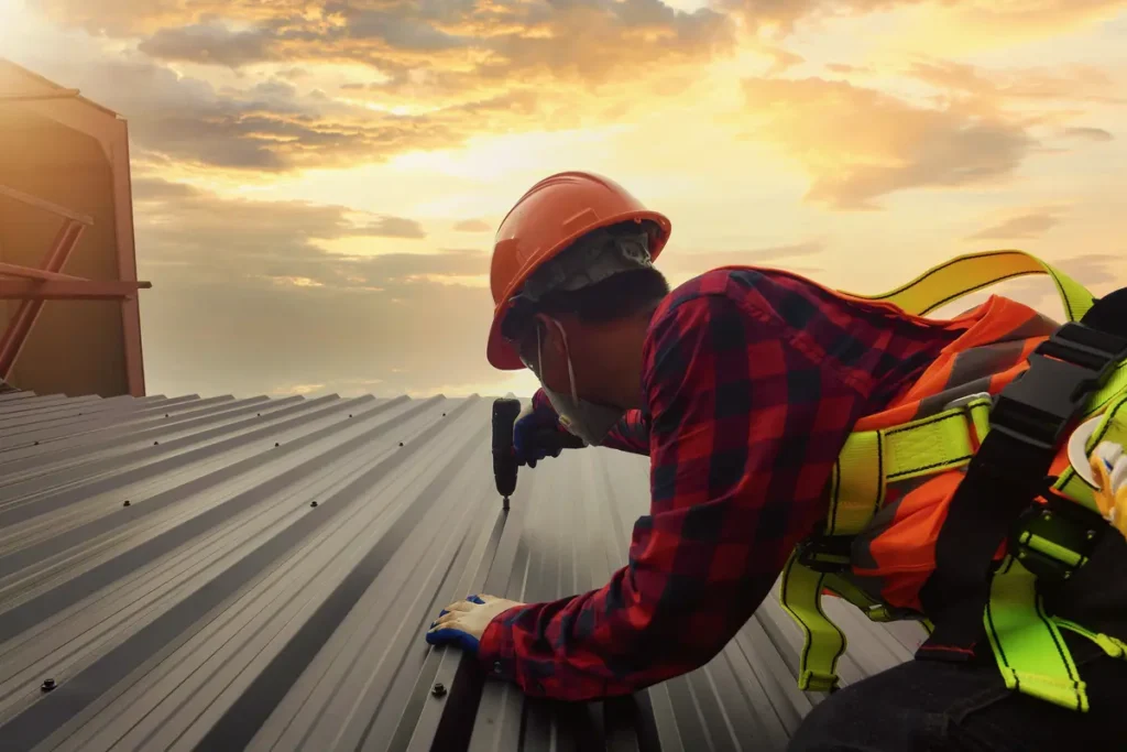 A construction worker in a safety harness and helmet drills into a metal roof panel at sunset. The sky is a mix of clouds and warm colors, creating a dramatic backdrop. The worker is wearing a red and black checkered shirt.