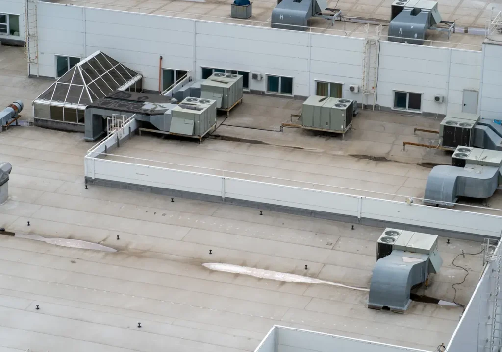 A flat commercial rooftop with several HVAC units and vents. The roof includes a small triangular glass structure. The surrounding tall white walls have small windows and pipes attached.