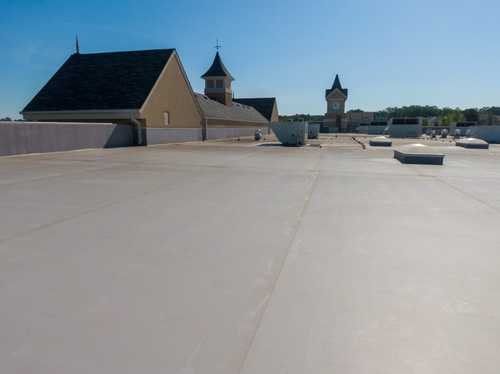 Flat rooftop with a smooth, light-colored surface under a clear sky. In the distance, there are buildings with pitched roofs and a clock tower. Ventilation units and skylights are scattered across the roof. Trees are visible on the horizon.