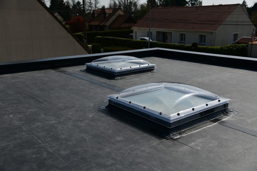 Flat rooftop with two dome-shaped skylights. The surrounding area includes neighboring houses and trees, indicating a residential setting. The skylights have a clear, modern design, letting light into the building below.