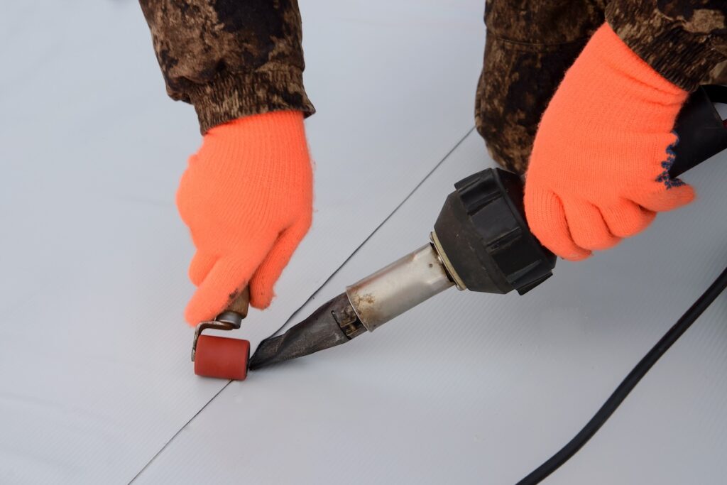 Person wearing bright orange gloves uses a heat gun and small roller to weld or seal a seam on a gray surface. Focus is on hands and tools, suggesting a precise task.