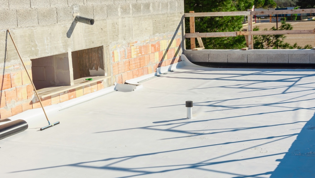 A flat rooftop under construction with white waterproofing material partially installed. Bricks and concrete are visible on the unfinished walls. A broom is propped against the wall, and a pipe emerges from the roof surface. Shadows are cast across the area.