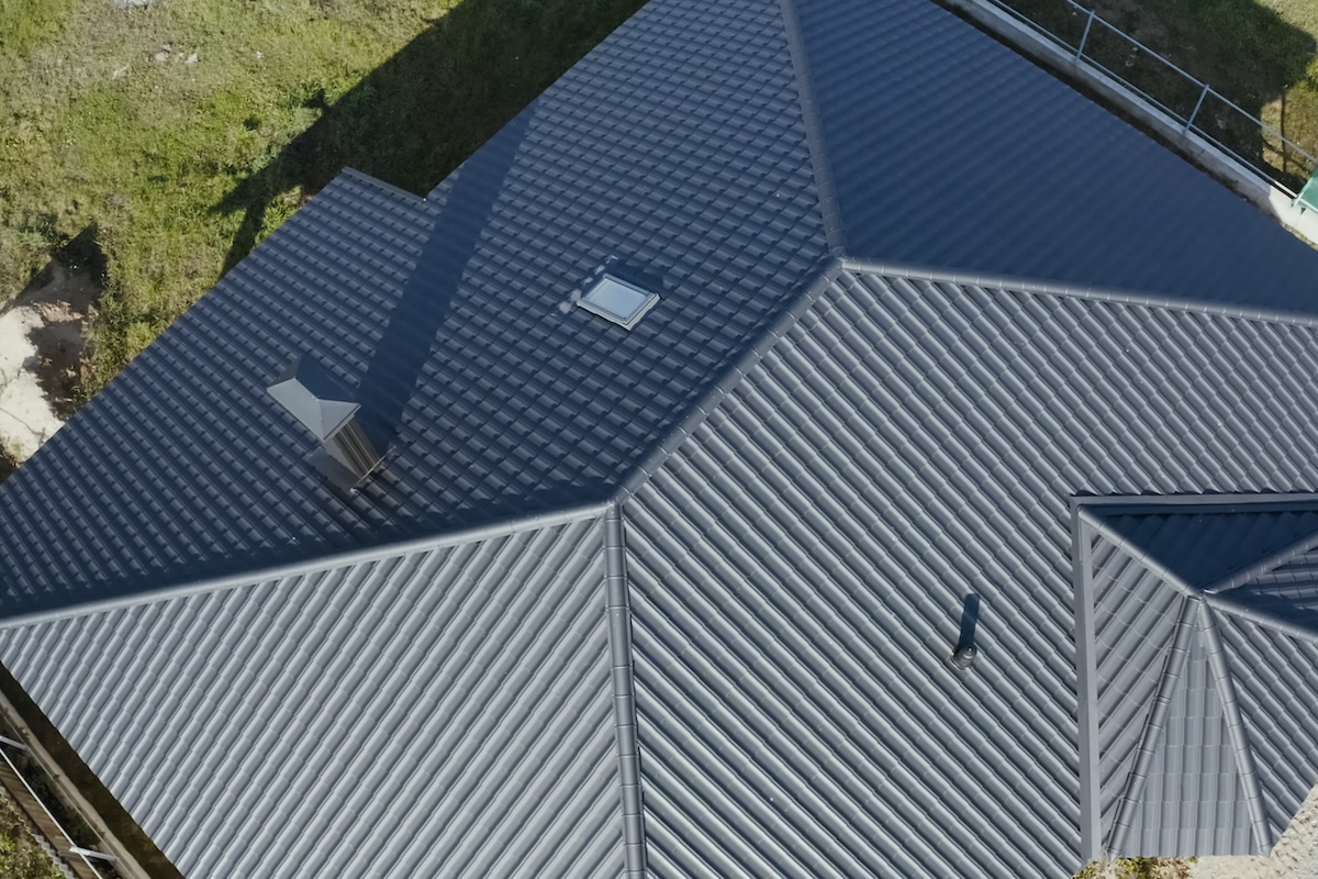 Aerial view of a building's dark gray, textured metal roof with multiple peaks and two vents. The roof also features a small skylight. Green grass is visible around the building's edges.