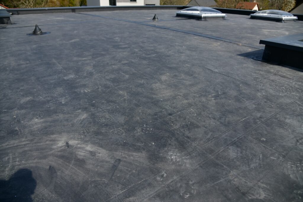 A flat rooftop with a smooth, dark surface featuring skylights and a few small protrusions. The background shows a glimpse of a building and trees under a clear sky.