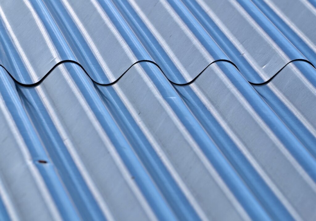 A close-up view of a corrugated metal sheet showcasing its wavy pattern and metallic texture. The sheet has a reflective, silver surface and is highlighted by natural light, creating a play of shadows and highlights along the ridges and grooves.