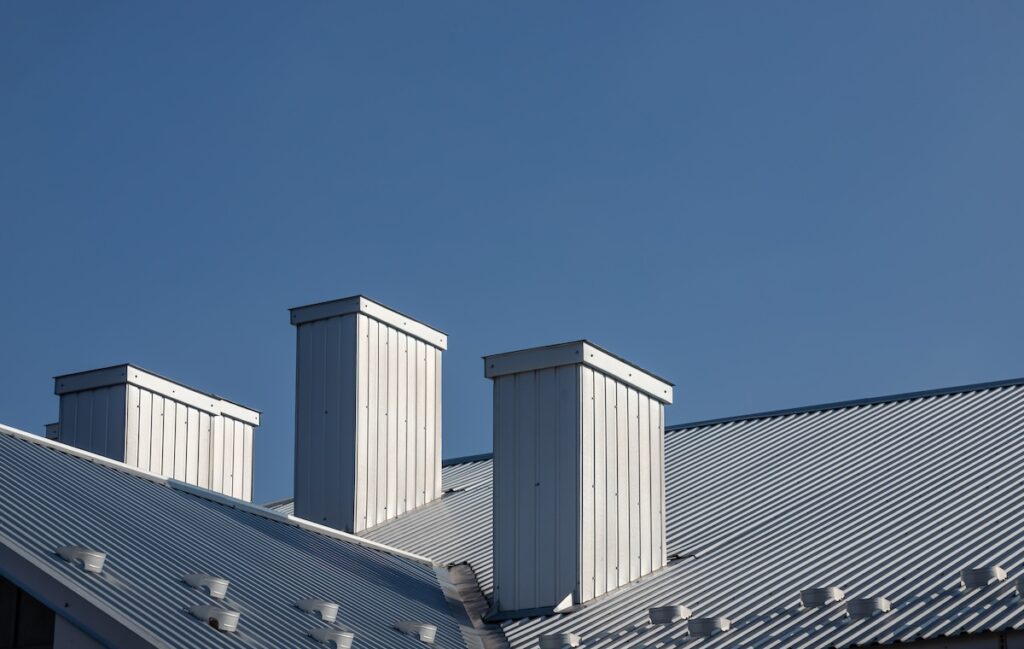A metal roof with three rectangular white chimneys against a clear blue sky. The roof's corrugated texture is visible, and sunlight highlights the chimneys, casting subtle shadows.