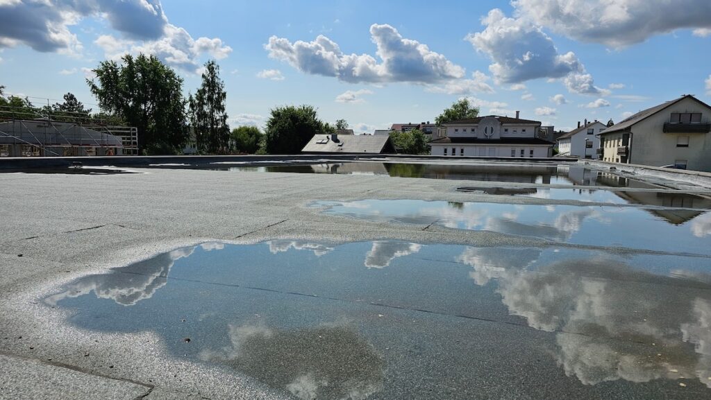 Rooftop with reflective puddles of water showing clouds and blue sky. Buildings and trees are visible at the edges under a sunny sky with scattered clouds.