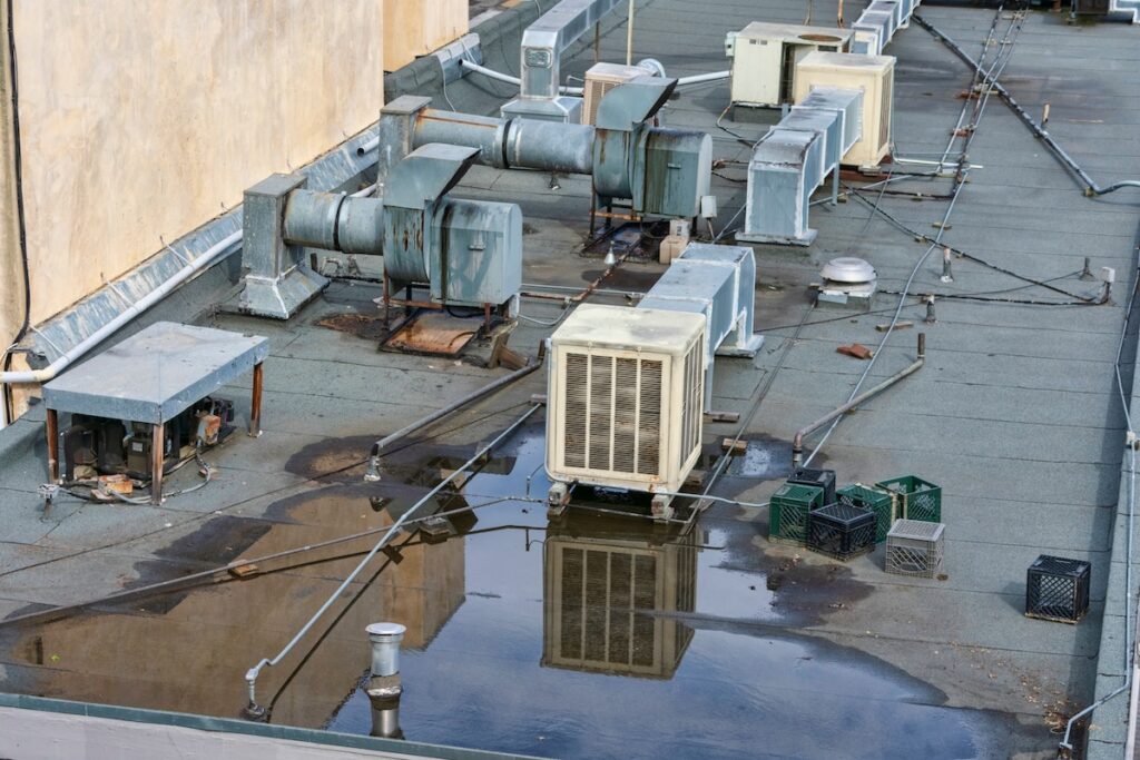 Rooftop view of an industrial building with multiple HVAC units and metal ductwork. A large puddle of water reflects the equipment. Several milk crates and wires are scattered around the roof, which has a flat, weathered surface.
