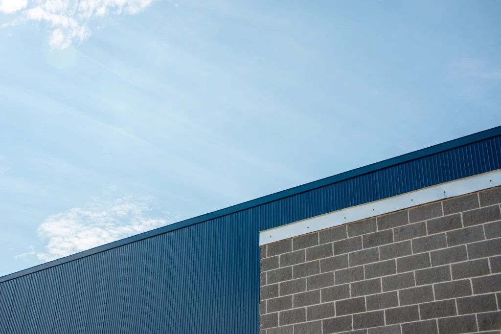 A modern building with a gray cinder block wall and blue metal facade, featuring flat roof flashing, is shown against a clear sky 