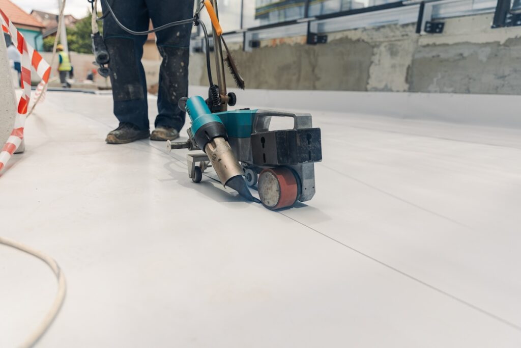 A person operates a blue and silver automatic welding machine on a white surface, smoothing and sealing material joints. Tape marking is visible on the left. The background shows unfinished walls and another person holding equipment.