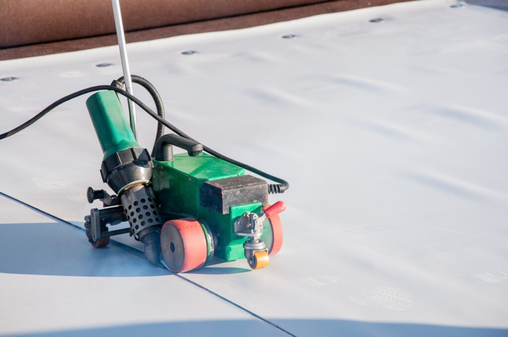 A green and black automatic welding machine on a flat roofing membrane. It has red wheels and a nozzle for sealing seams as it moves across the white surface.