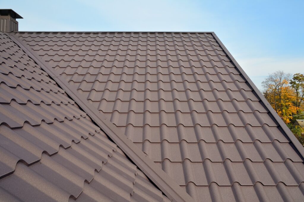 A close-up view of a brown metal roof with a ridge and interlocking panels, under a clear blue sky. The roof design features a patterned, wavy texture, and trees can be seen in the distant background.