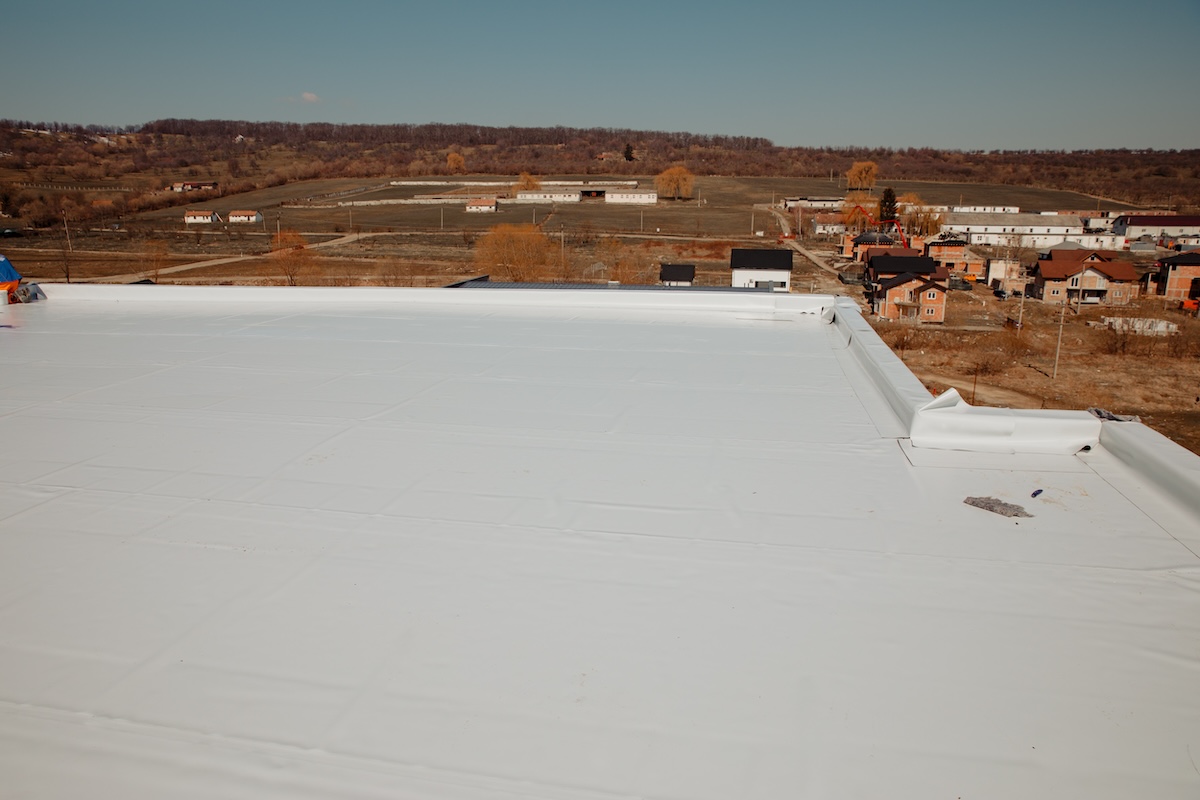 A flat white rooftop overlooking a rural landscape with houses, fields, and trees under a clear blue sky. In the distance, small buildings and a forested area are visible.