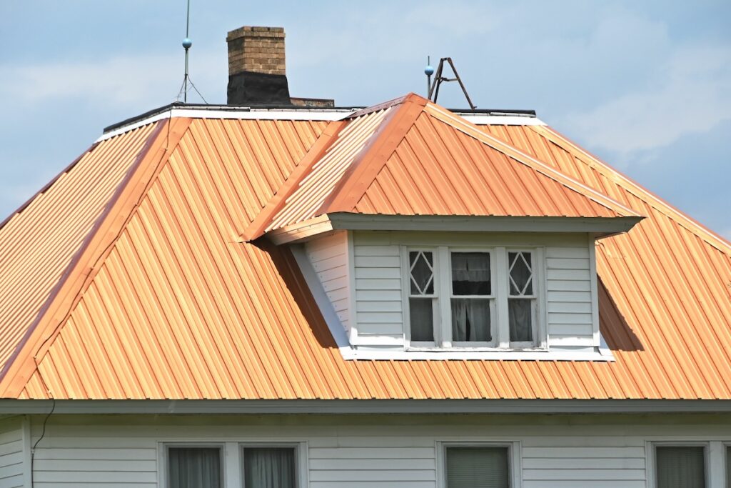 Close-up of a house with a copper-colored, metal hip roof featuring a white dormer. The dormer has a double window with decorative crossbars. A brick chimney is visible behind the dormer, and the sky is partly cloudy.