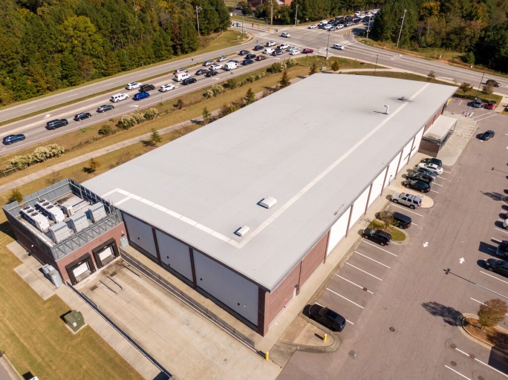 A large rectangular warehouse with a gray roof and brick siding, surrounded by a parking lot with several cars. The building is near a busy road with multiple cars. Trees and greenery are visible in the background.