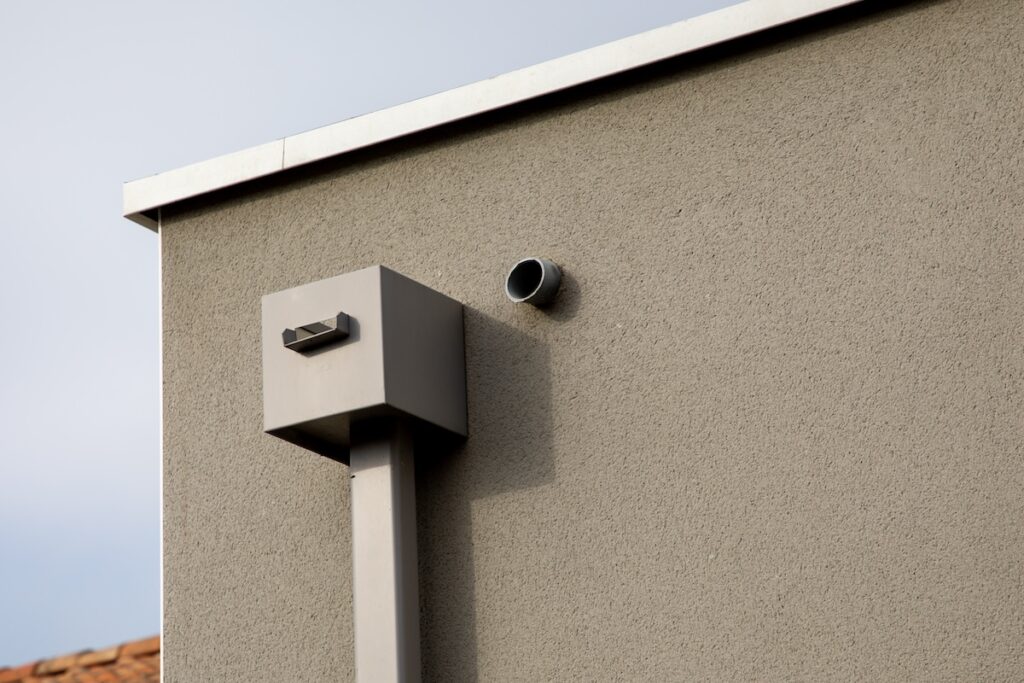 Exterior view of a building with a light brown stucco wall. Features include a protruding rectangular structure attached to a vertical pipe and a small circular vent further up the wall. The sky is clear in the background.