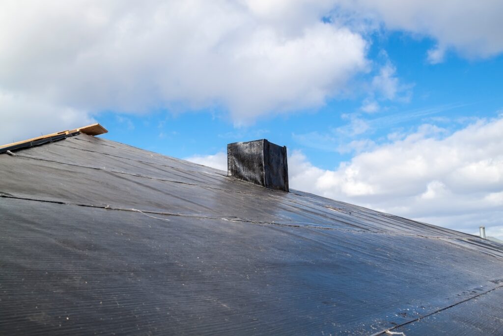 A sloped roof covered with black tar paper under a partly cloudy sky. A square black chimney-like structure projects from the roof's surface. A piece of wood is visible near the roof's edge on the left.