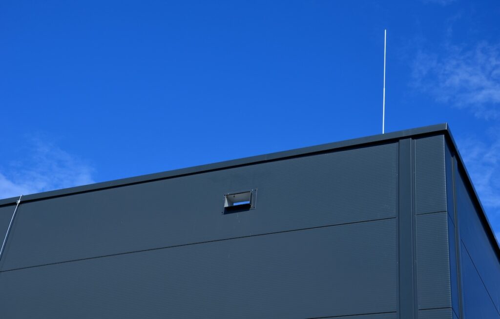 The top corner of a modern building with a dark exterior is shown against a clear blue sky. A small rectangular window is visible, along with a slender antenna protruding from the roof's edge.