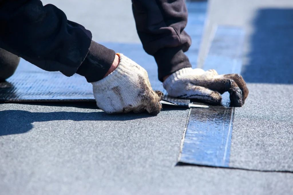 A person wearing gloves is installing roofing material, carefully aligning black sheets with metallic strips on a flat surface. The focus is on their hands and the materials being used.