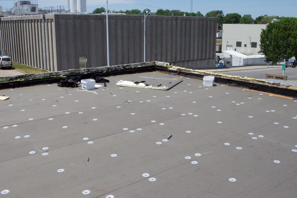 Flat rooftop with visible sheet metal and screws. Two white buckets, debris, and a large rectangular panel are scattered. In the background are industrial buildings, parked cars, and trees under a clear sky.