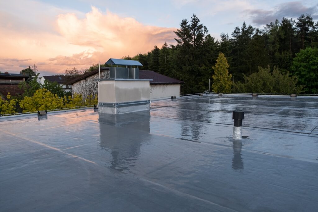 A flat rooftop with a shiny, reflective surface, likely after rain. There's a chimney-like structure and a few vents. Surrounding the roof are trees and a house. The sky is partially cloudy with a warm sunset glow.