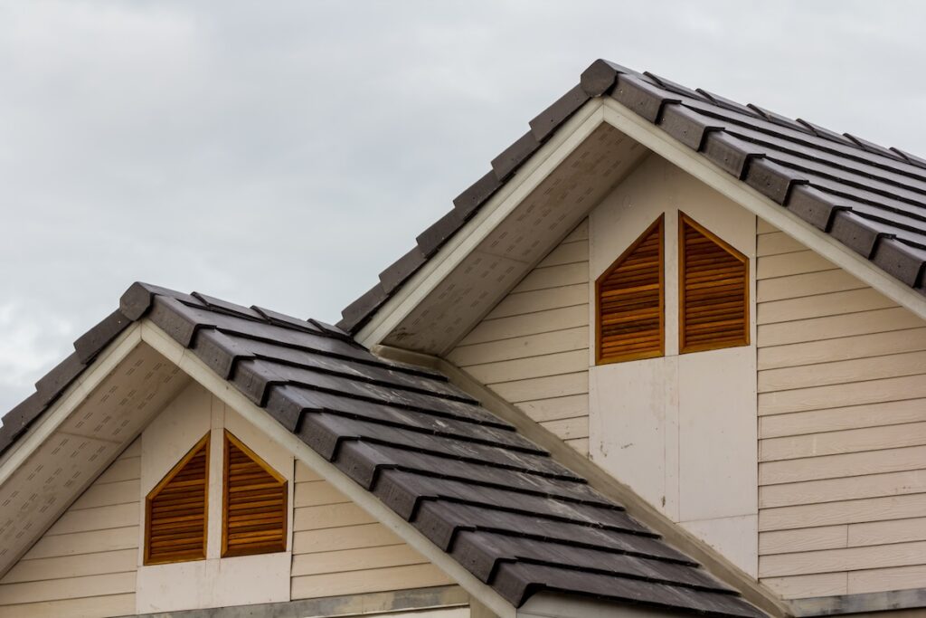 Close-up of two house gables with dark gray tiled roofs and light beige siding. Each gable features a triangular wooden vent with horizontal slats. The sky in the background is overcast.