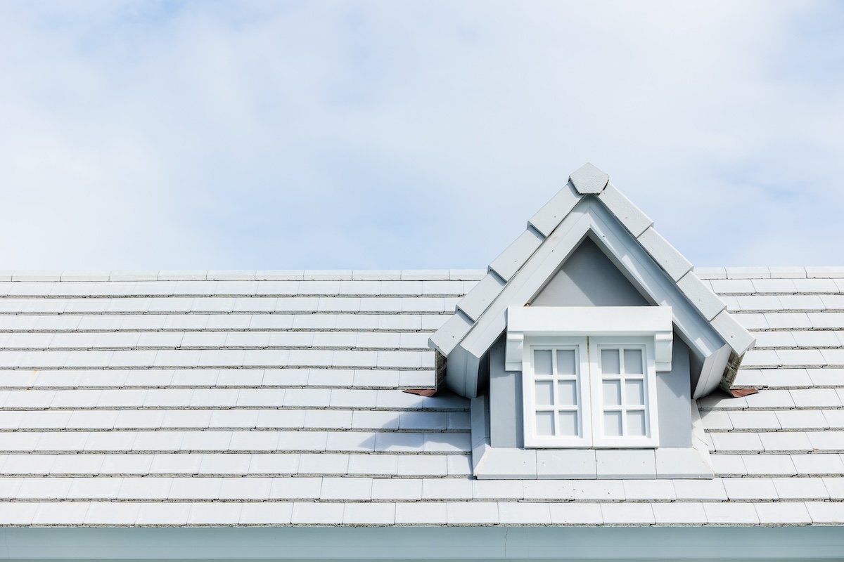 A light gray, tiled roof with a small dormer window in the center against a cloudy sky. The dormer has a triangular top and is painted white, matching the roof's clean and uniform appearance.
