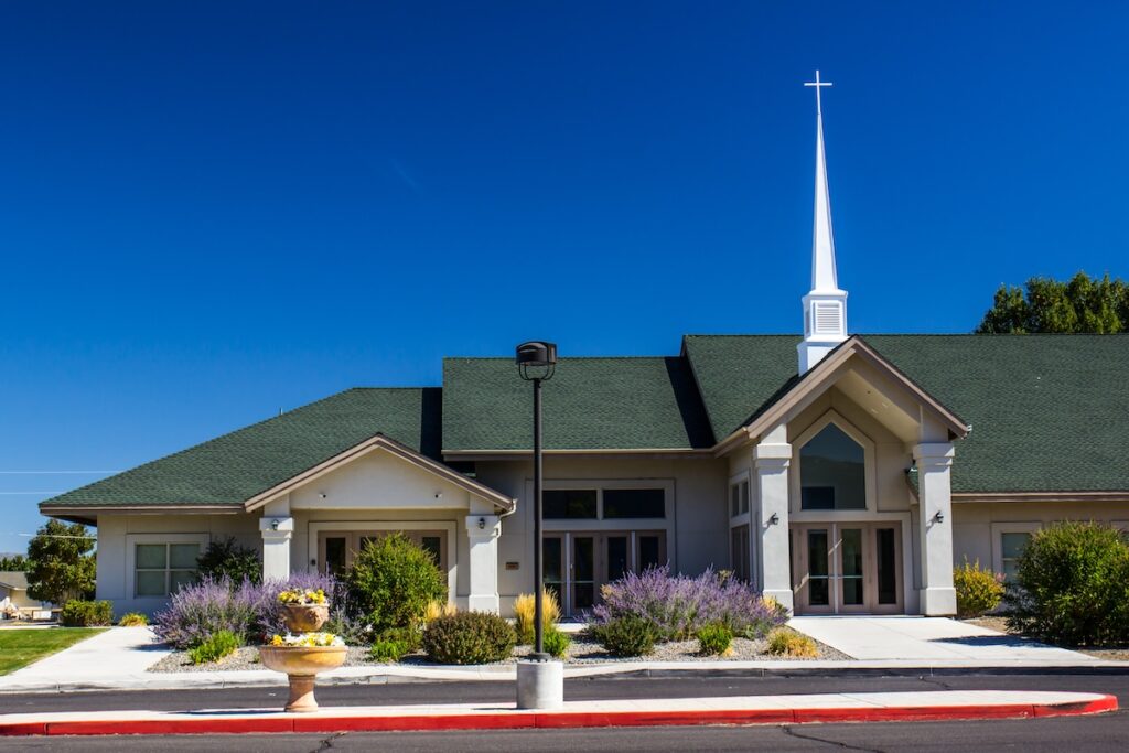 A modern church with a green roof and a tall white steeple against a clear blue sky. The building is surrounded by neatly landscaped gardens with purple and yellow flowers, a pathway, and a red curb in the foreground.