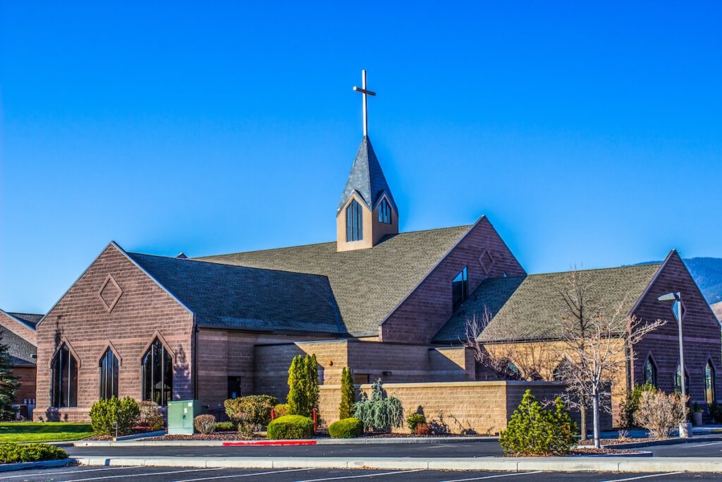 A large, brown, wooden church with a tall steeple topped by a cross under a clear blue sky. The building has several sections with peaked roofs and is surrounded by shrubs and a paved road in the foreground.
