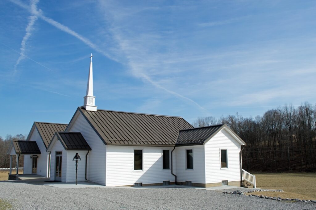 A small white church with a steeple and dark roof stands on a gravel path. It's surrounded by bare trees, and a clear blue sky is above.