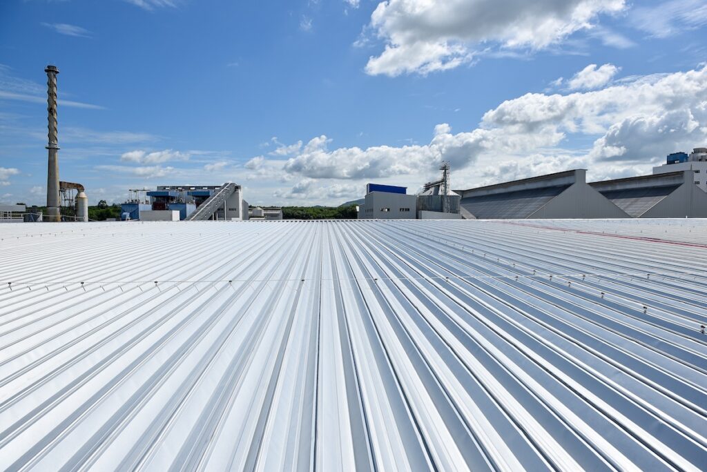 View of a large, corrugated metal rooftop under a bright blue sky with scattered clouds. Industrial structures, including a chimney, are visible in the background, suggesting a factory or warehouse environment.