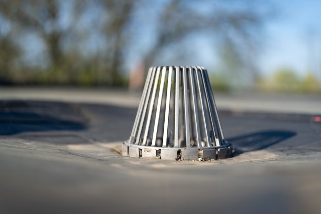 A close-up of a rooftop drain with a metal guard that looks like a cage. The background is blurred, showing hints of trees and clear blue sky.