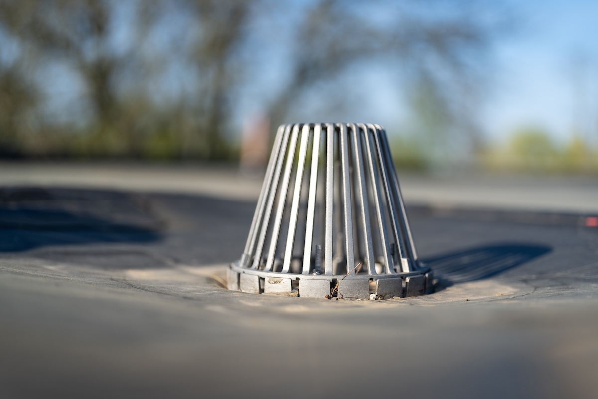 A close-up of a rooftop drain with a metal guard that looks like a cage. The background is blurred, showing hints of trees and clear blue sky.
