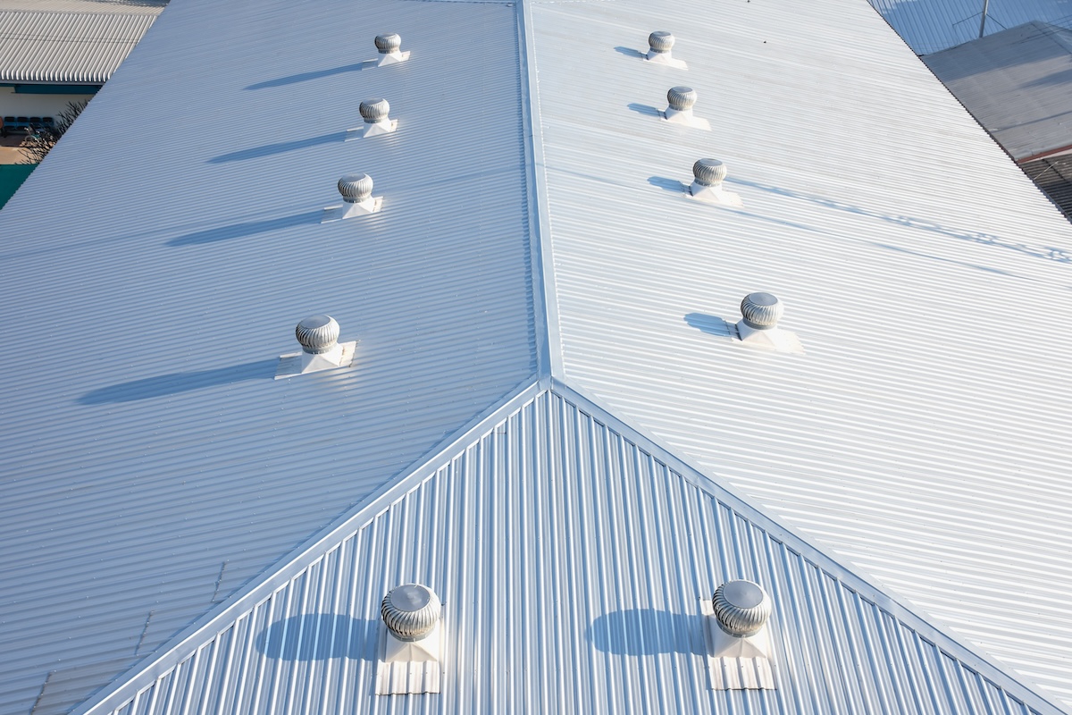 Aerial view of a large, white industrial roof with multiple evenly spaced ventilation turbines along the ridgeline. The roof is corrugated, and shadows of the turbines are visible on the surface.