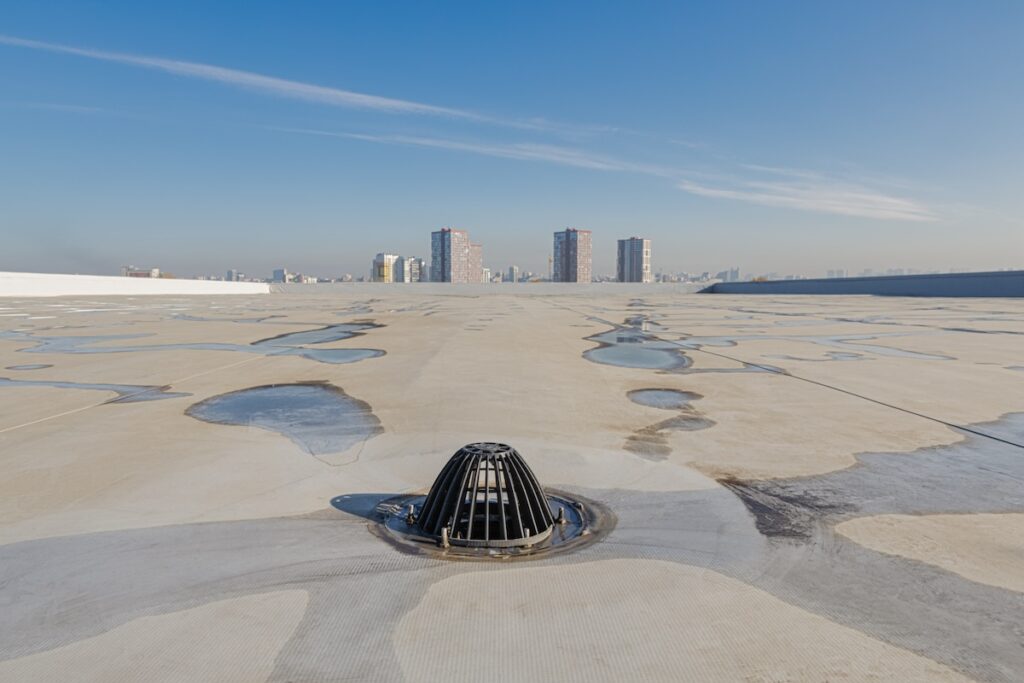 Flat rooftop with a central drainage grate surrounded by drying water patches. The background shows a clear blue sky and a city skyline with several mid-rise buildings.