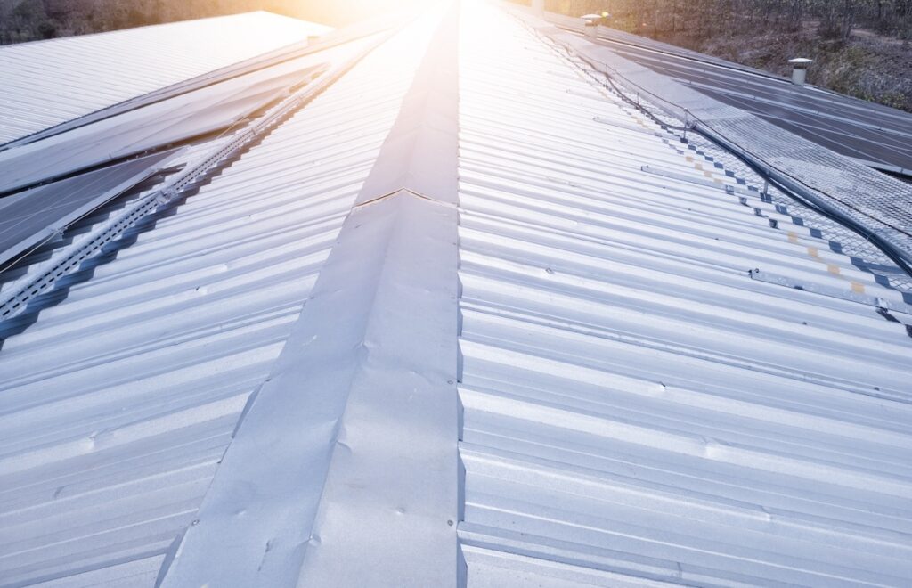 View of a corrugated metal roof reflecting sunlight, extending towards the horizon. Sunlight creates a bright, glowing effect, while trees and buildings are faintly visible in the background.