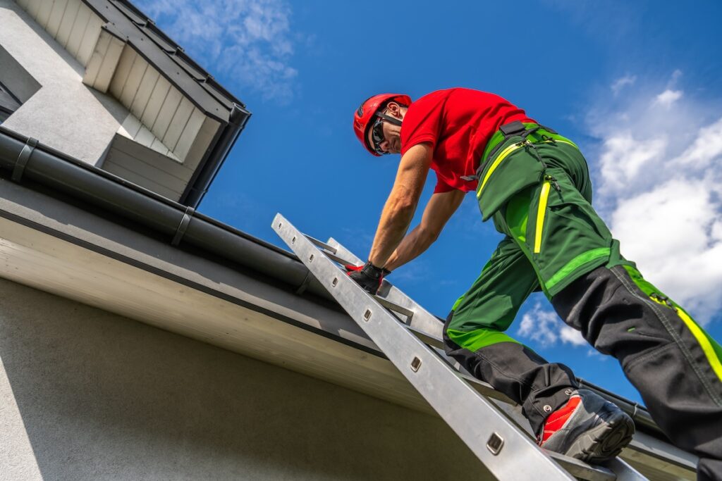 A worker in a red shirt and helmet climbs a metal ladder to reach the roof of a building. He is wearing green and black protective gear and gloves. The sky is clear and blue, and the building's roof and gutters are visible.
