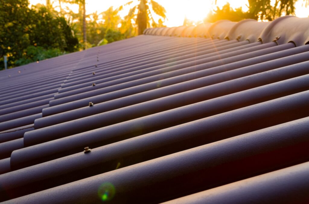 Close-up of a tiled rooftop at sunset, with sunlight glowing in the background. The roof tiles are shiny with rows leading to the horizon. Trees and a warm sky are visible in the distance, casting a serene atmosphere.