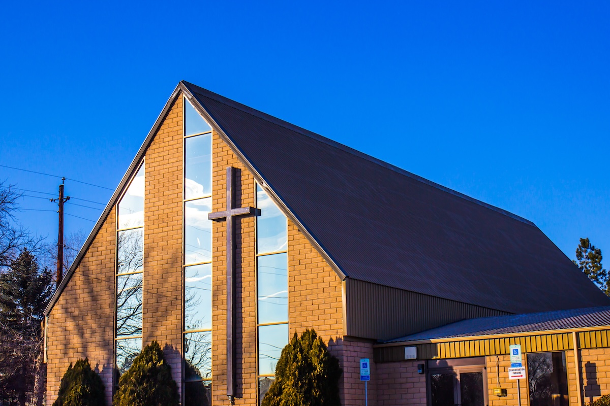 A modern church with a tall, sloped roof and large glass windows reflecting the blue sky. A cross is mounted on the facade. The building is surrounded by trees, and a clear, sunny day illuminates the scene.