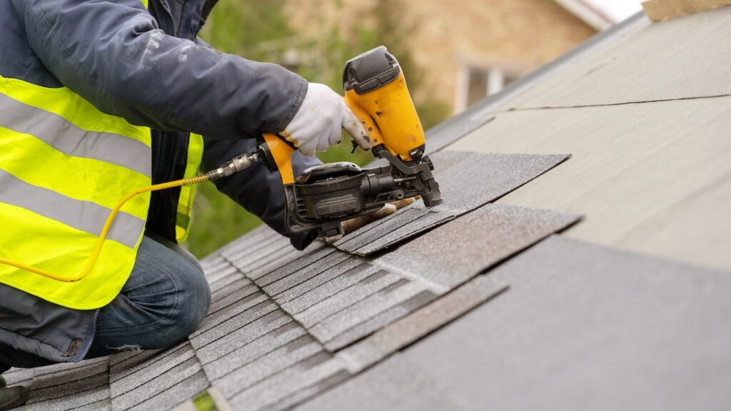 Person in a safety vest and gloves using a pneumatic nail gun to install gray asphalt shingles on a sloped roof. The scene suggests active roofing work on a clear day.