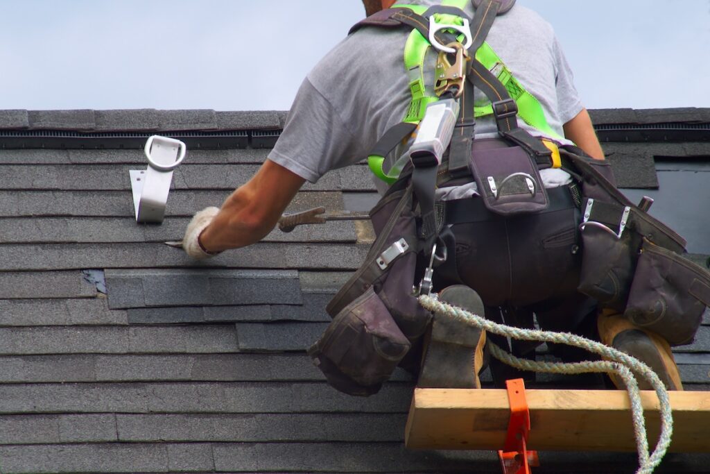 A worker wearing a safety harness is repairing a shingled roof. They have a tool belt with various tools and are using a utility knife to adjust the shingles. A safety rope and ladder are visible, ensuring the worker's safety.