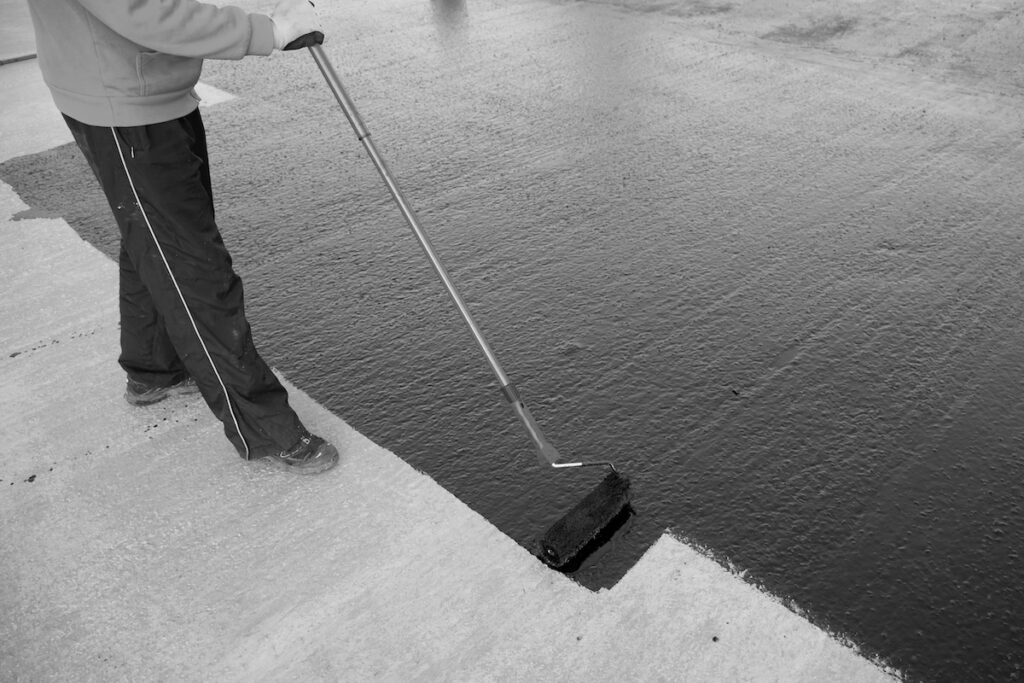 A person uses a paint roller attached to a long handle to apply modified bitumen roofing solution to commercial roof