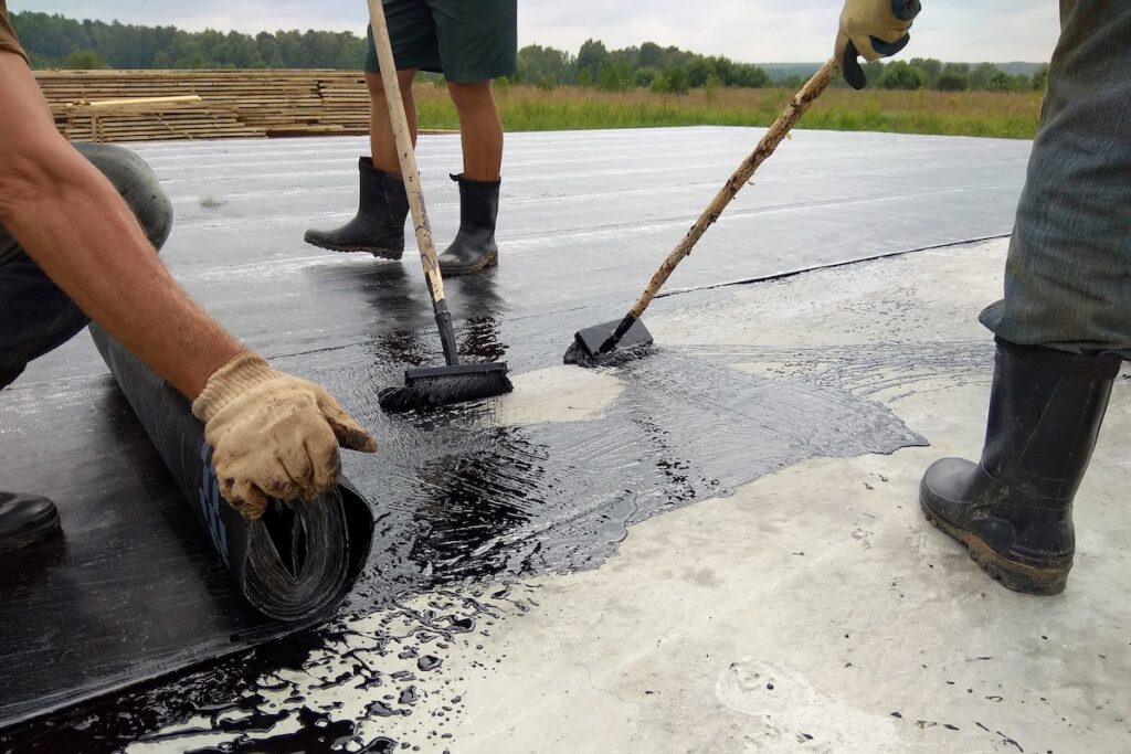 Three workers in boots and gloves apply black waterproofing material to a concrete surface using rollers and brushes, with one person unrolling a sheet